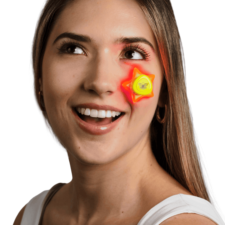 A smiling woman demonstrating the use of a yellow, star-shaped Metamorphosis LED light therapy device under her eye, which emits red light for targeted anti-aging, wrinkle reduction, and skin rejuvenation treatments.
