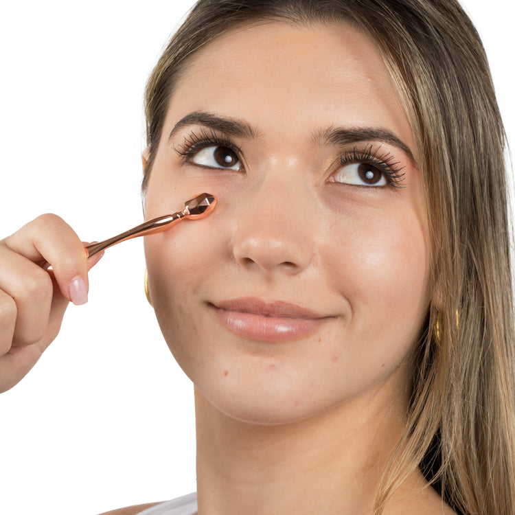 A Metamorphosis smiling model using a small, copper-toned mini eye roller on her under-eye area to reduce puffiness, promote circulation, and apply eye cream.