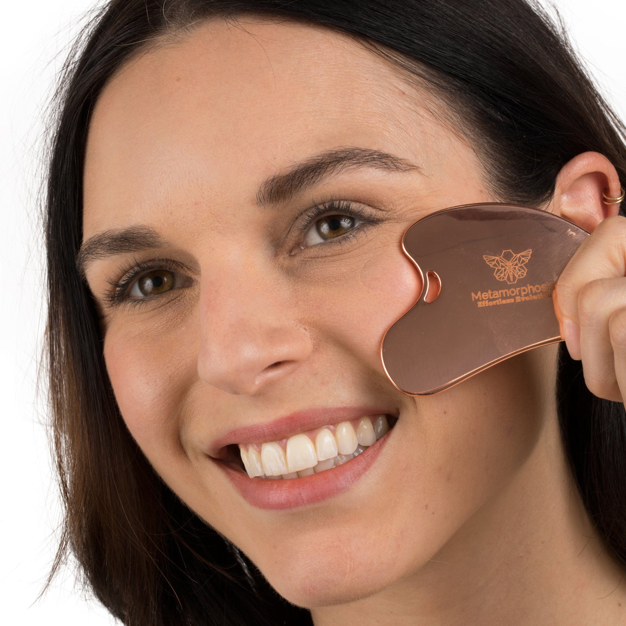 A smiling model using a copper-toned Metamorphosis Gua Sha facial tool on her cheek, promoting facial sculpting, lifting, and contouring for a radiant complexion.