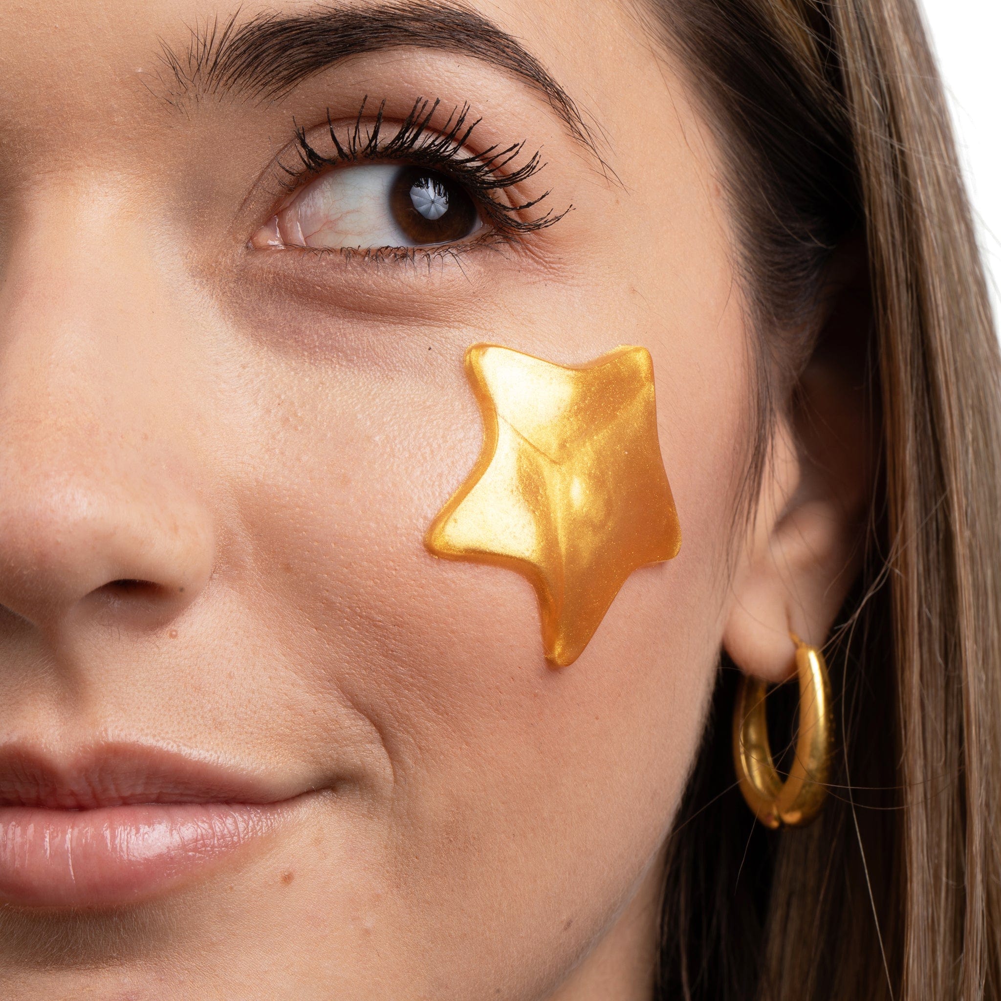 Metamorphosis A close-up shot of a woman with her eyes open, slightly smiling, wearing a gold, star-shaped hydrogel eye mask or pimple patch on her cheek for hydration and skin treatment.