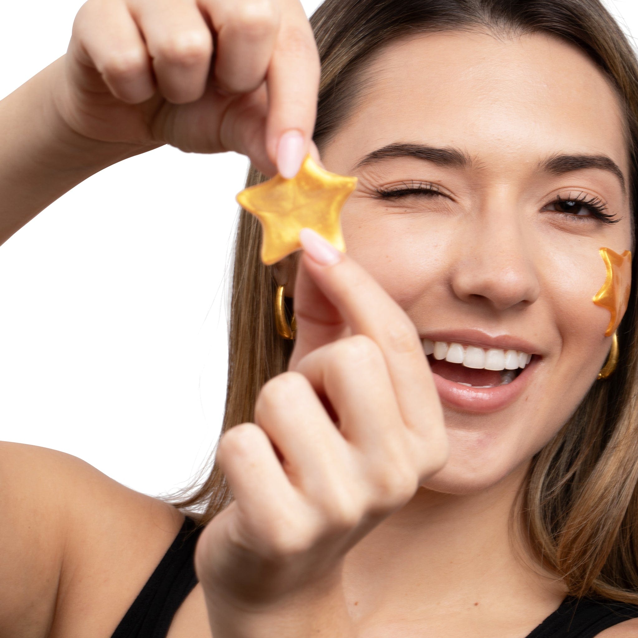 A smiling woman winking and holding a gold, star-shaped Metamorphosis hydrogel eye patch, showcasing the SuperStar Pi product for under-eye hydration, brightening, and skin recovery.