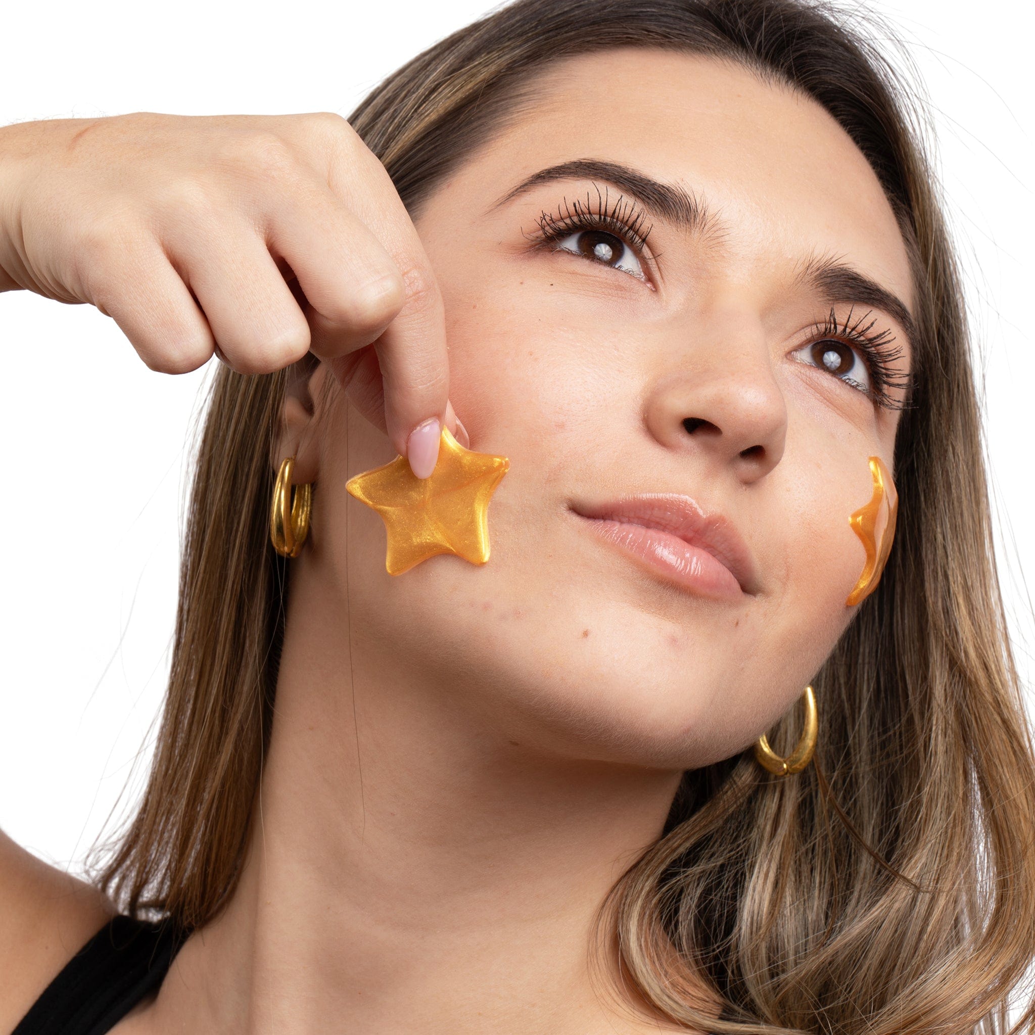 A model applying a gold, star-shaped hydrogel eye mask or pimple patch to her cheek, demonstrating the application of a skincare product for hydration or acne treatment.